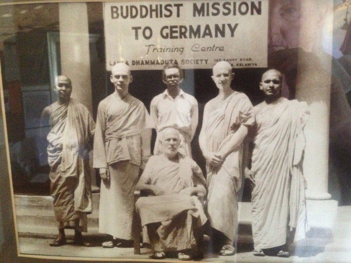 Das Bild zeigt sechs Männer (fünf stehend, einer sitzend) vor einem Gebäude, zwischen dessen Säulen ein Banner mit der Aufschrift "Buddhist Mission to Germany" gespannt ist.