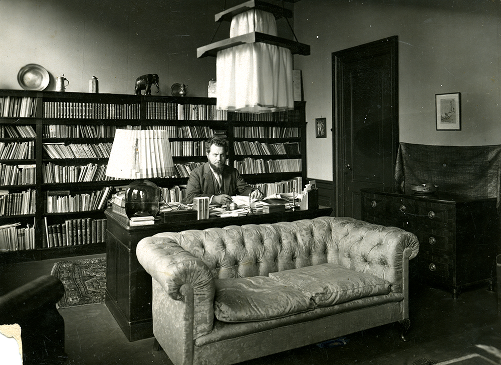 A black and white photograph of Ernst Kraus sitting at his desk in front of an impressive book shelf.