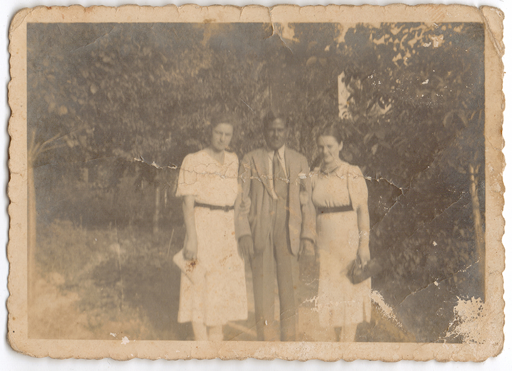 An aged black and white photograph showing Sakhawat Hussein Khan posing with two women surrounded by trees.