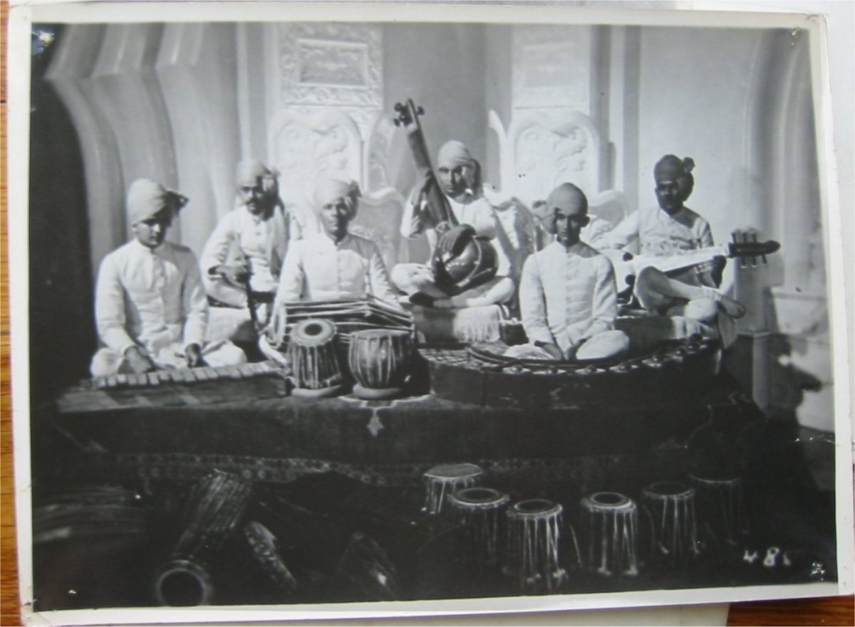 A black and white image showing six musicians on stage, some holding musical instruments prominent in South Asian classical music.