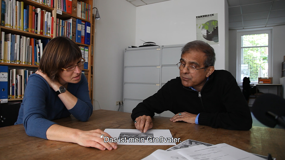 A still from a film showing Irfan Khan seated in what appears to be a library, conversing with a person whilst pointing at the print out of an image lying on the table in front of them. The moment is subtitled with the words "Das ist mein Großvater" [German for "This is my grandfather."