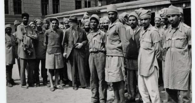 A group of around 20-30 POWs are standing together with the visitor Dr. Exchaquetin what appears to be a courtyard in front of a larger building.