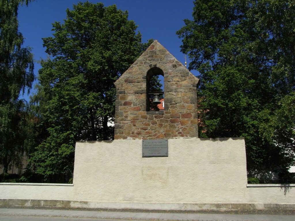 Überreste eines Gebäudes mit einigen Bäumen im Hintergrund. Besondere Merkmale der Ruine sind eine Glocke sowie eine Gedenktafel.