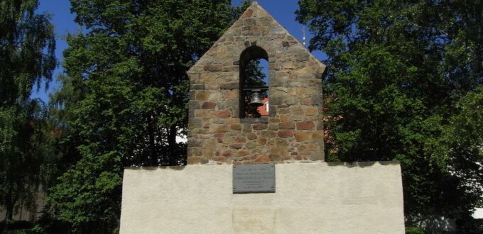 Überreste eines Gebäudes mit einigen Bäumen im Hintergrund. Besondere Merkmale der Ruine sind eine Glocke sowie eine Gedenktafel.