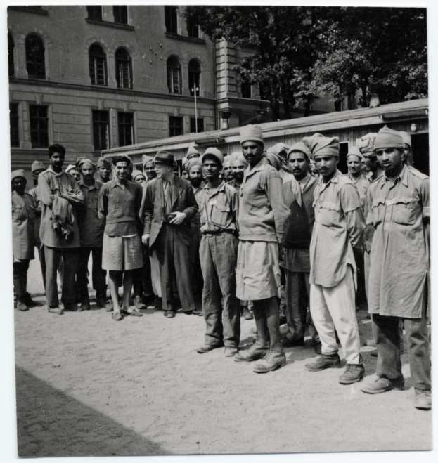 A group of around 20-30 POWs are standing together with the visitor Dr. Exchaquetin what appears to be a courtyard in front of a larger building.