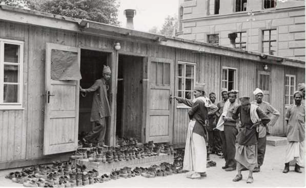 A group of about 8 POWs are standing outside of the small hutments which contain the prayer rooms. One POW is standing inside the entrance of one of the prayer rooms, leaning on the doorframe. The door is surrounded by a large number of shoes, neatly arranged in pairs.