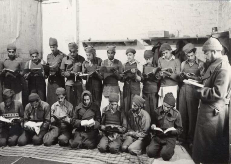 A group of Indian POWs are in a room reading from the Quran, 11 of them standing, 7 sitting.