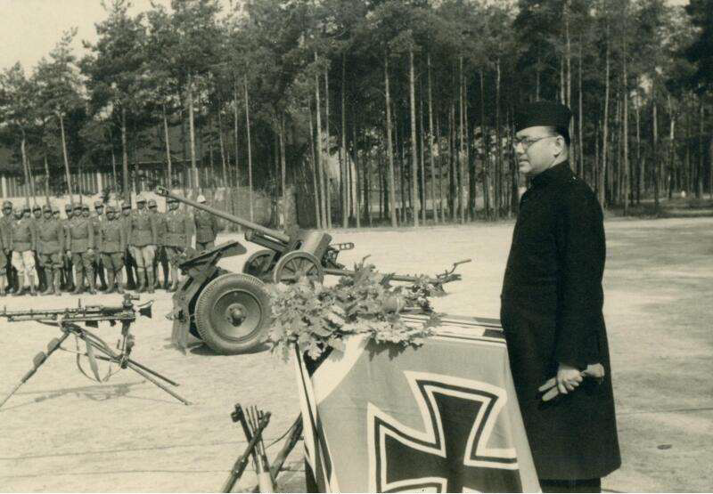 Netaji is standing in the foreground looking to the left, in front of him there is a flag with a large Iron Cross on it. Midground one can see a stationary machine gun and an anti-aircraft-gun. In the background there is a group of Indian POWs in soldiers' uniforms in front of a row of trees.