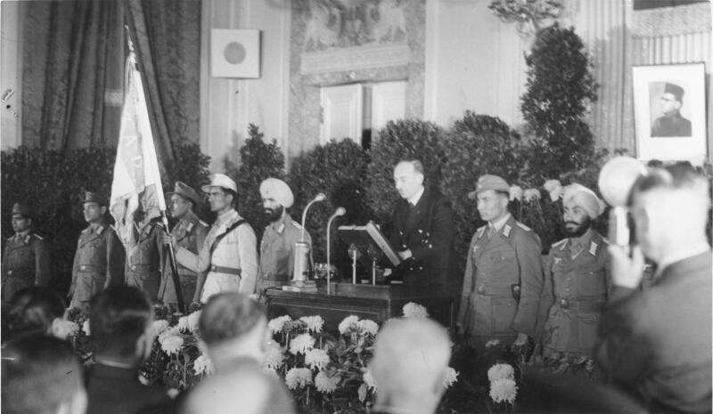A German official is adressing an audience inside a festive hall. To his left and right once can see a few Indian POWs in soldiers' uniforms.