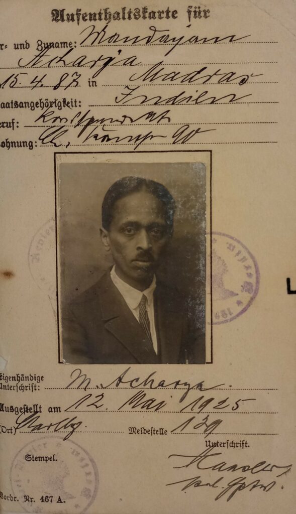 The image shows an archival source, particularly Acharya's German residence permit. It contains handwritten information about as well as a photograph of Acharya, wearting a suit and tie as well as a short mustache.