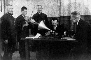 In this black-and-white photograph, Schünemann and Stumpf can be seen sitting at a table. They are operating the recording equipment. On the left side of the picture are three Tartar musicians, one of them playing a kind of violin or viola in front of the recording funnel.