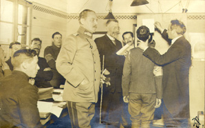 The image is a photograph showing Wilhelm Doegen and Alois Brandl in a room with eight other men. Doegel, standing, holds one of the men by the scruff of the neck and points his head into the speaking tube of the recording device. Brandl is standing next to him, another man - probably the recording technician - behind the equipment, a third next to Brandl - from his uniform probably a camp supervisor. The remaining men are sitting at the edge, presumably they are camp inmates.