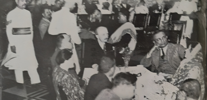 On this picture you can see a gathering of the Bangiya Jarman Vidya Samsad. There are three tables with 36 guests in total, predominantly dressed in suits and sarees. Among them are well-known people such as the Samsad's founder, Benoy Kumar Sarkar and the mayor of Calcutta. In the left corner, there are two waiters, dressed in traditional white garments.