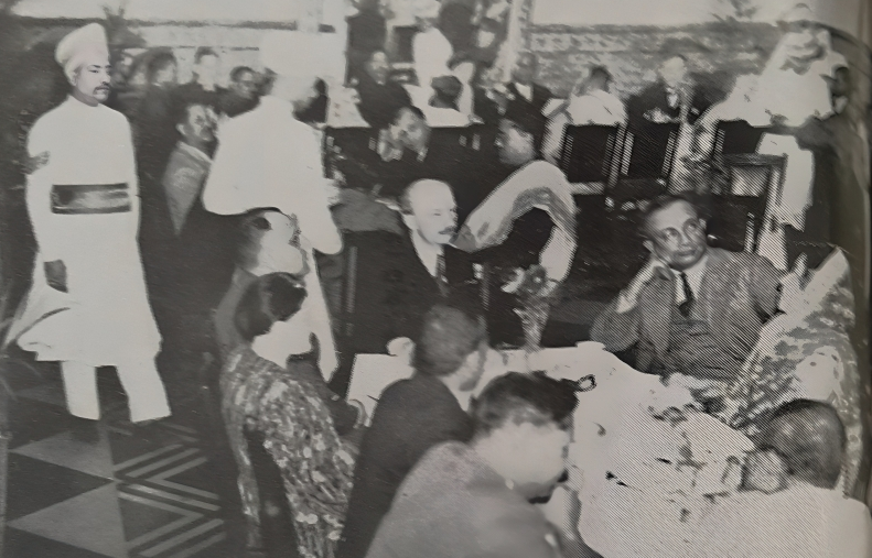 This photograph shows a gathering of the Bangiya Jarman Vidya Samsad. There are three tables with 36 guests in total, predominantly dressed in suits and sarees. Among them are well-known people such as the Samsad's founder, Benoy Kumar Sarkar and the mayor of Calcutta. In the left corner, there are two waiters, dressed in traditional white garments. 