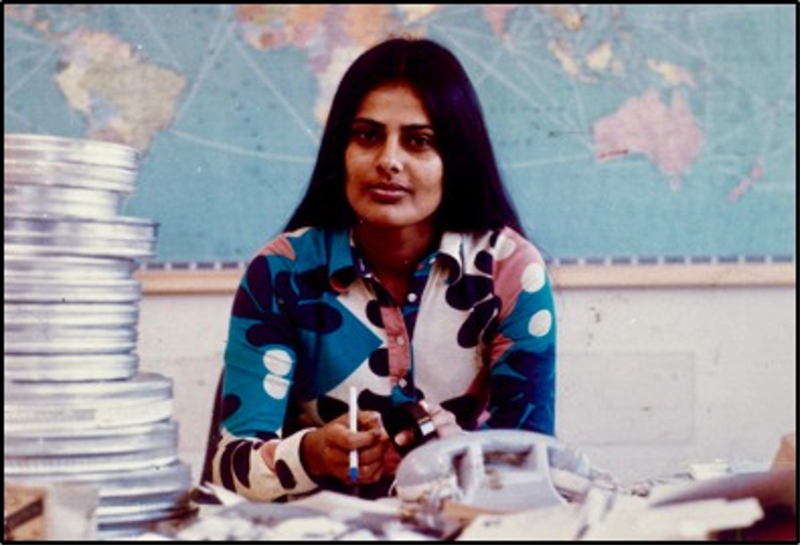 Das Foto zeigt ein Porträt von Navina Sundaram. Sie sitzt an einem Schreibtisch, hinter ihr ist eine Weltkarte zu sehen. Auf dem Schreibtisch gestapelt, links von ihr liegen Filmrollen, Navina Sundaram selbst trägt eine bunte Bluse im Stil der 70er-Jahre. Ihre langen Haare trägt sie offen und in der Mitte gescheitelt.