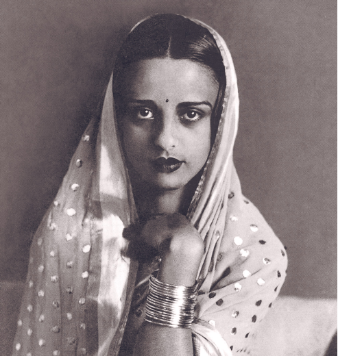 Amrita Sher-Gil blickt auf diesem schwarz-weiß Foto direkt in die Kamera. Sie trägt einen gepunkteten Sari und mehrere, wahrscheinlich goldene, Armreifen. Ihr Haar ist in der Mitte gescheitelt.