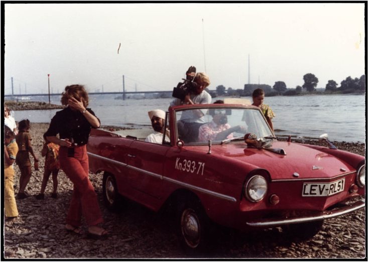 Ein Einzelbild aus der Reportage zeigt Darshan Singh in seinem roten Wagen am Ufer des Rheins. Links neben dem Auto steht eine Frau (vielleicht Sundaram) und einige Kinder. Hinten im Wagen filmt ein Kameramann das Geschehen.
