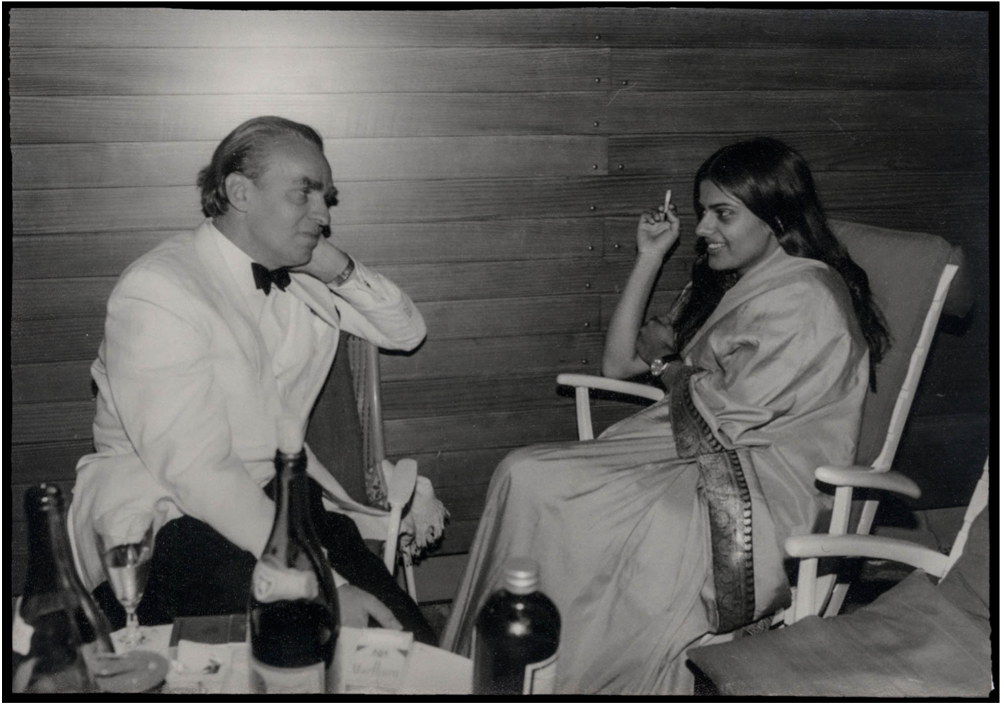 Ein schwarz-weiß Foto, das Sundaram und Bode auf Stühlen sitzend beim Genuss von Wein und Zigaretten zeigt. Sundaram trägt einen Sari, Bode ein weißes Jacket mit dunkler Fliege.