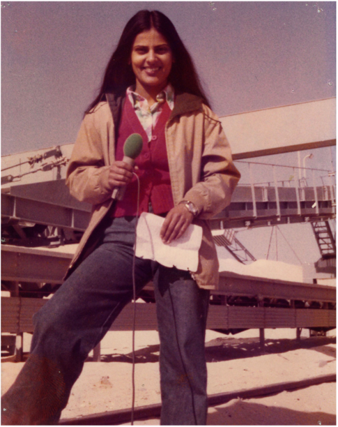 Ein altes Farbfoto zeigt Navina Sundaram an einem sonnigen Tag stehend vor einigen Förderbändern. Ihr Haar ist in der Mitte gescheitelt und sie lächelt. Sie trägt eine rote Bluse, eine beige Jacke, und eine dunkle Hose. In der rechten Hand hält sie ein Mikrofon, in der linken ihre Notizen.