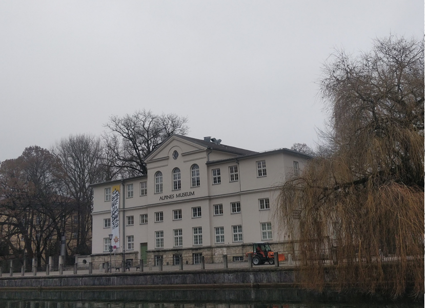 The picture shows the front of the Alpine Museum, with the entrance facing the Isarkanal. It is a cloudy winter’s day, and the surrounding trees are bare.